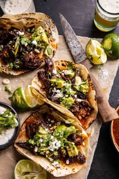 three tacos with various toppings on a cutting board next to two glasses of beer