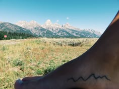 a person with a tattoo on their foot sitting in the grass near mountains and trees