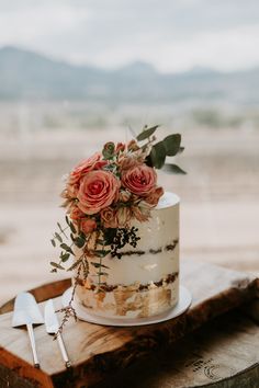 a white and gold wedding cake with pink flowers on top sitting on a wooden slab