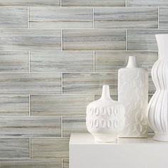 three white vases sitting on top of a shelf next to each other in front of a gray tile wall