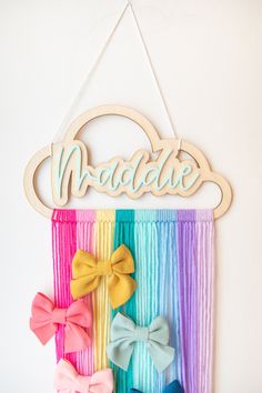 a rainbow wall hanging with wooden name and three bow clips on it's side