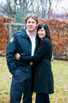 a man and woman standing next to each other in front of some trees with leaves on the ground