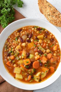 a white bowl filled with vegetable soup next to a piece of bread and parsley