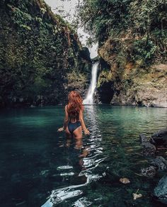 a woman in a swimsuit standing in the water near a waterfall and looking up into the sky