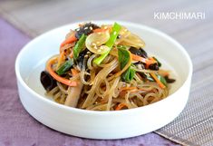 a white bowl filled with noodles and vegetables on top of a table next to a purple cloth