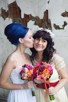 two brides are kissing each other with flowers in front of an old peeling wall