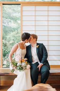 a bride and groom sitting on a bench kissing each other in front of a window