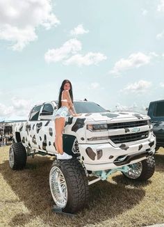 a woman standing on the hood of a white truck with cow print paint job and big tires