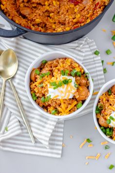 two bowls filled with chili and rice next to silver spoons on a white table