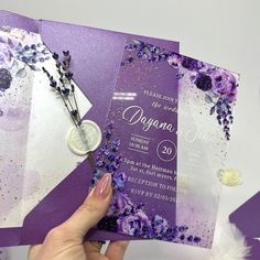 a purple and white wedding card with flowers on it, being held up by a woman's hand