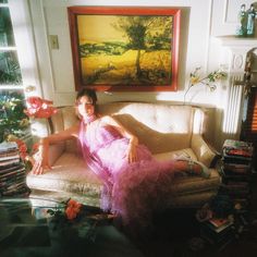 a woman in a pink dress sitting on a couch next to a painting and bookshelf