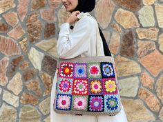 a woman wearing a hijab and carrying a crocheted bag in front of a stone wall