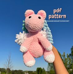 a hand holding a pink and white crocheted stuffed animal in the air with trees in the background