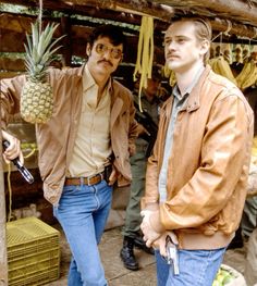 two men standing next to each other near bananas and pineapples