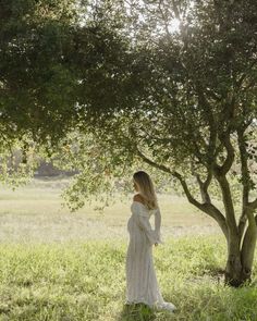 a woman in a white dress is standing under a tree and looking at the ground