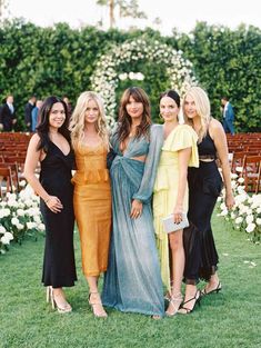 four women standing next to each other in front of an outdoor ceremony area with white flowers and greenery