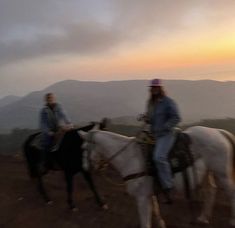 two people riding horses on top of a hill at sunset or dawn with mountains in the background