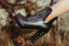 A person's leg wearing the Western Edge Boutique's HIGH DEMAND *LUX black leather ankle boot is extended towards the camera. The high-quality boot boasts intricate white stitching and metal studs on the chunky heel. A small triangle tattoo adorns the person's ankle, while a cactus can be seen in the blurry background. Candle Room, Curvy Dress, It Girl, Leather Silver, Hat Hairstyles, Clothing And Shoes, Bag Accessories, Shoe Accessories, Lounge Wear