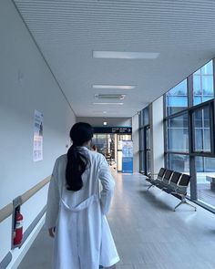 a woman in white coat walking down a hallway next to a bench and large windows