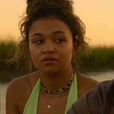 a woman in a green shirt and necklace looking at the camera with an orange sky behind her