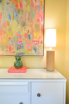 an abstract painting hangs above a white dresser with two lamps on it and a green vase sitting on top