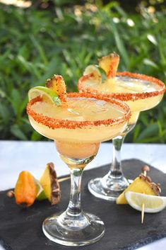 two glasses filled with drinks sitting on top of a black tray next to lemon wedges