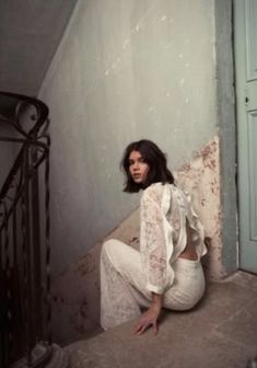 a woman is sitting on the ground in white pants and a lace top, leaning against a wall