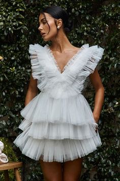 a woman wearing a white dress standing in front of a bush with her hands on her hips