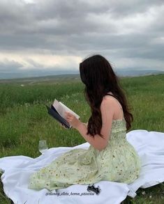 a woman sitting on top of a white blanket reading a book in a green field