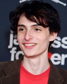 a young man wearing a red shirt and brown blazer smiles at the camera while standing in front of a black background