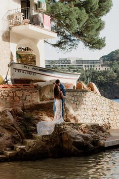 a bride and groom are standing on the rocks by the water in front of a boat