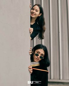 two young women are peeking out from behind a wall, one is holding onto the pole