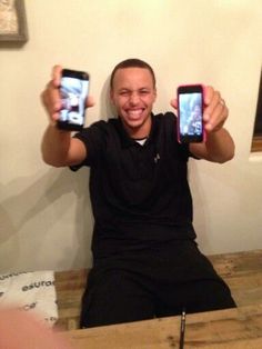 a man holding up two cell phones in front of his face while sitting on a table