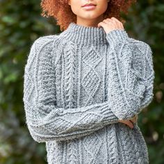 a woman with red curly hair wearing a gray cabled sweater and jeans standing in front of trees