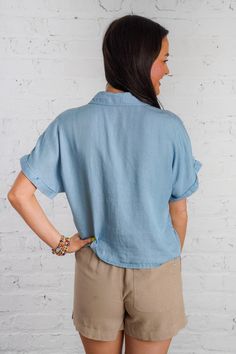 Ride the waves of style with our Drifting Tides Top. This blue chambray top features a button down front, a tie detail, and a front pocket, perfect for a casual yet chic look. The short cuffed drop sleeves and collared neckline add a touch of charm, while the soft chambray material provides ultimate comfort. Pair with our Sand Dune Shorts and Sunbeam Raffia Mules to complete the look! This top is lightweight, non-sheer, and has a loose boxy fit. 100% Tencel Brand- Be Cool Model is 5'5" and is we Washed Blue Chambray Button-up Top, Denim Beach Top With Short Sleeves, Beach Denim Short Sleeve Top, Blue Top With Pockets And Collared Neckline, Blue Collared Top With Pockets, Blue Collared Top With Roll-up Sleeves, Summer Collared Tops With Buttoned Pockets, Collared Denim Blue Blouse With Pockets, Relaxed Fit Light Blue Blouse With Pockets