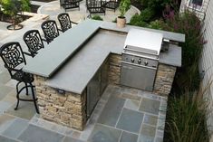 an outdoor kitchen with grill and chairs around the outside space, surrounded by stone patio furniture