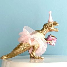 a gold dinosaur figurine in a tutu skirt and party hat holding a present