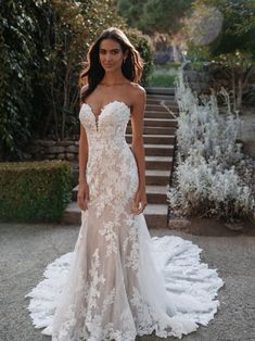 a woman in a white wedding dress standing on the ground next to some stairs and bushes