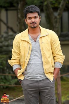 a man standing in front of a building wearing a yellow jacket and gray shirt with his hands in his pockets