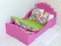 a pink doll bed with green pillows and flowers on the pillowcase, in front of a floral wallpapered background