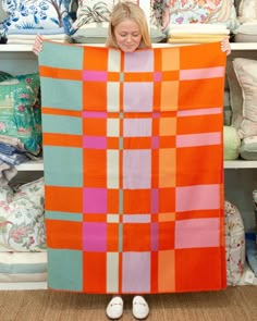 a woman is holding up an orange and pink blanket in front of some pillows on shelves