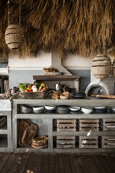 an outdoor kitchen with lots of plates and bowls on the counter, hanging from thatched roof