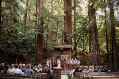 an outdoor ceremony in the woods with lots of people sitting on benches and standing around