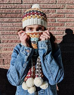 a woman wearing a knitted hat and scarf covers her face while standing in front of a brick wall