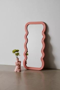 a vase with flowers and a pink mirror on a table next to it, all in front of a white wall