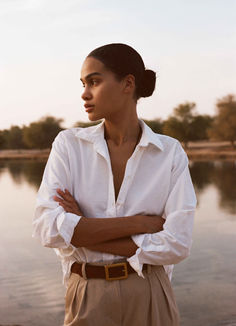 a woman standing in front of a body of water with her arms crossed and looking off to the side