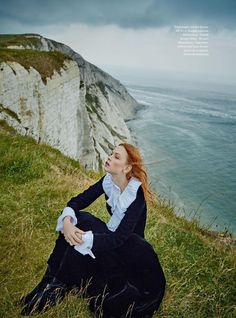 a woman sitting on top of a grass covered hillside next to the ocean and cliffs