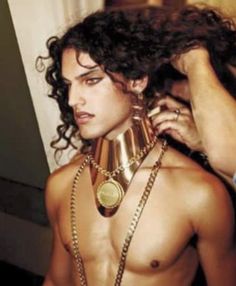 a shirtless man wearing a gold collar and chain around his neck, while another person combs his hair