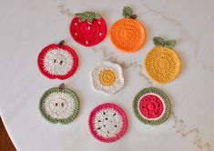 crocheted fruit coasters are arranged on a marble countertop, including an apple, orange, and kiwi