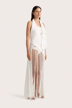 A halter neck dress with draped fabric over the shoulders and a semi-fitted bodice. Cut from 100% cotton jersey, it features an asymmetrical hemline detailed with handwoven macrame and a fringe skirt that falls down around the ankles. Style the Bebe Bikini Bottom underneath and add a simple leather sandal for a unique approach to occasionwear. Asymmetrical Maxi Dress, Maxi Dress White, Halter Neck Dress, Maxi Dress Sale, Simple Leather, Fringe Skirt, White Maxi, Halterneck Dress, Draped Fabric